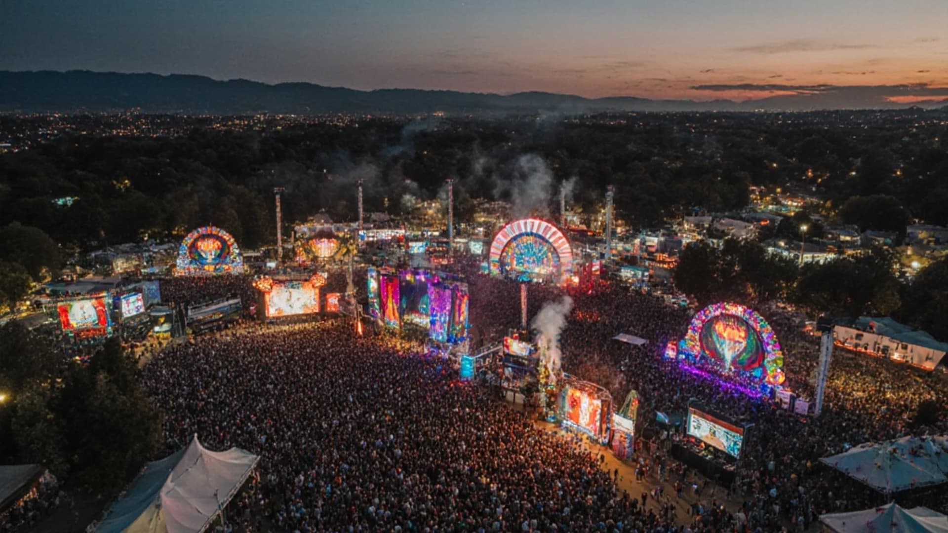 Twilight aerial photography capturing vibrant music festival with illuminated stages and thousands of attendees