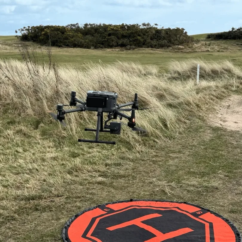 A drone lifting off from rough coastal grass in strong wind, with no buildings or people anywhere in sight — the kind of place an A3-only drone has to be flown