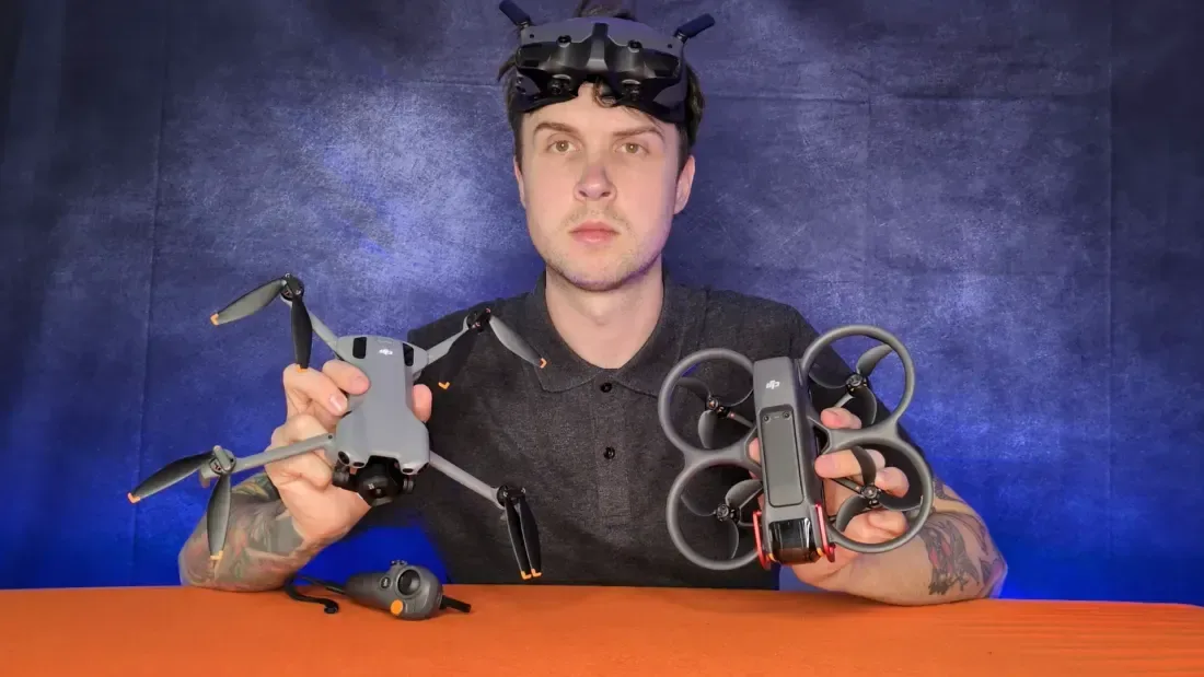 Peter Leslie with DJI FPV goggles pushed up on his forehead, holding a folding DJI camera drone in one hand and a caged DJI Avata 2 in the other, with an RC Motion controller on the table