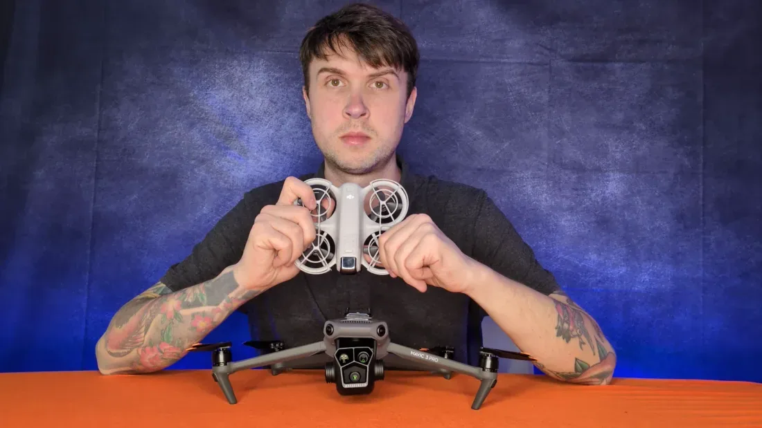 Peter Leslie holding a small caged DJI Neo 2 in both hands above a much larger DJI Mavic 3 Pro sitting on the table — the two ends of the DJI range in one frame