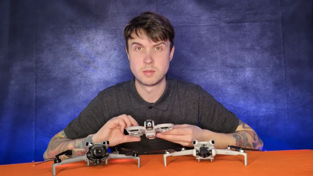 Peter Leslie lifting a caged DJI Neo 2 off the table between a DJI Mini 5 Pro on the left and a DJI Mini 4 Pro on the right — three sub-250-gram DJI drones together