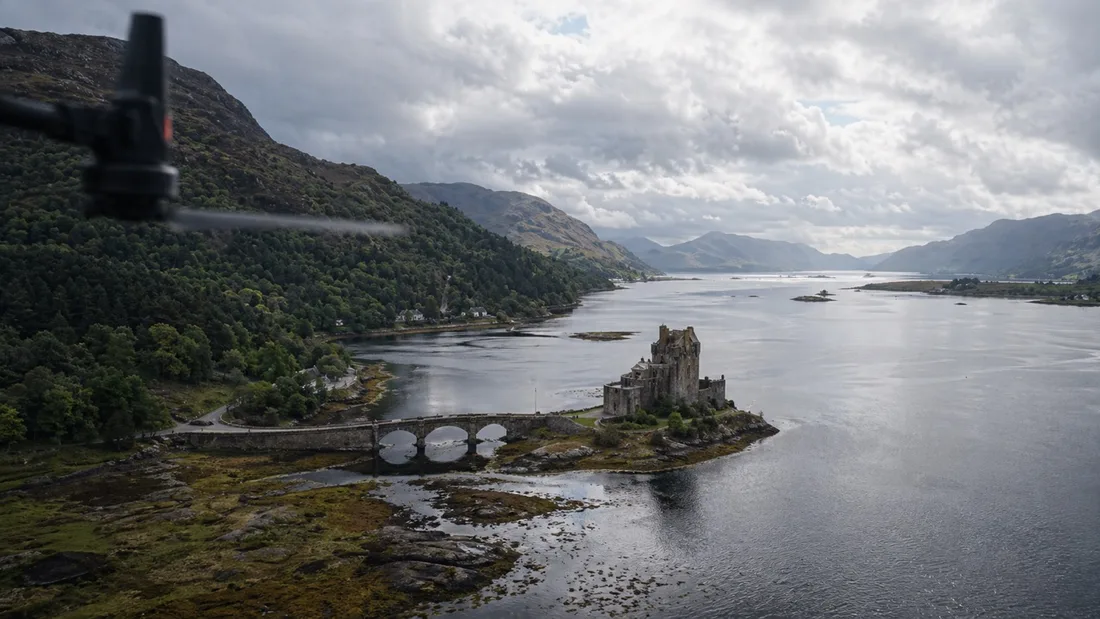 Aerial view along a Scottish sea loch with steep wooded hillsides falling to the water and a castle on a small island, a drone propeller blurred at the top of the frame