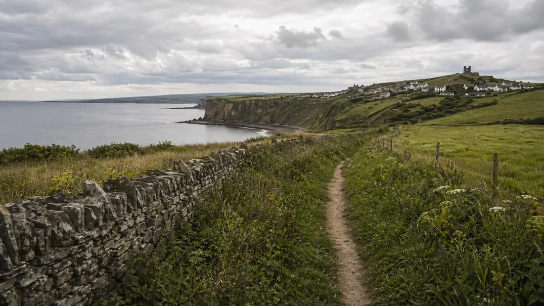 A narrow footpath running along a grassy clifftop above a bay, with a ruined tower on the headland ahead and a dry stone wall on the left