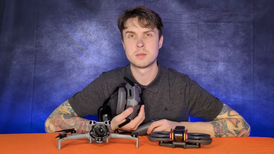 Peter Leslie holding a DJI RC 2 controller in one hand, with a DJI Mini 5 Pro and an FPV drone on the table in front of him