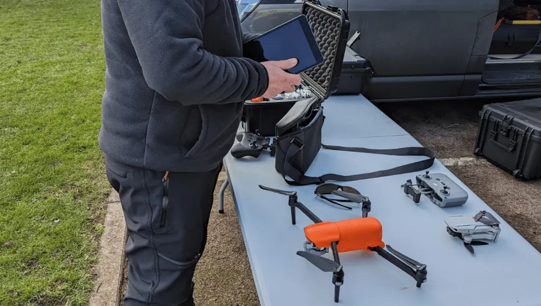 A full drone kit laid out on a table beside a van: a folded grey drone, an orange Autel drone, two controllers, a tablet and a hard case