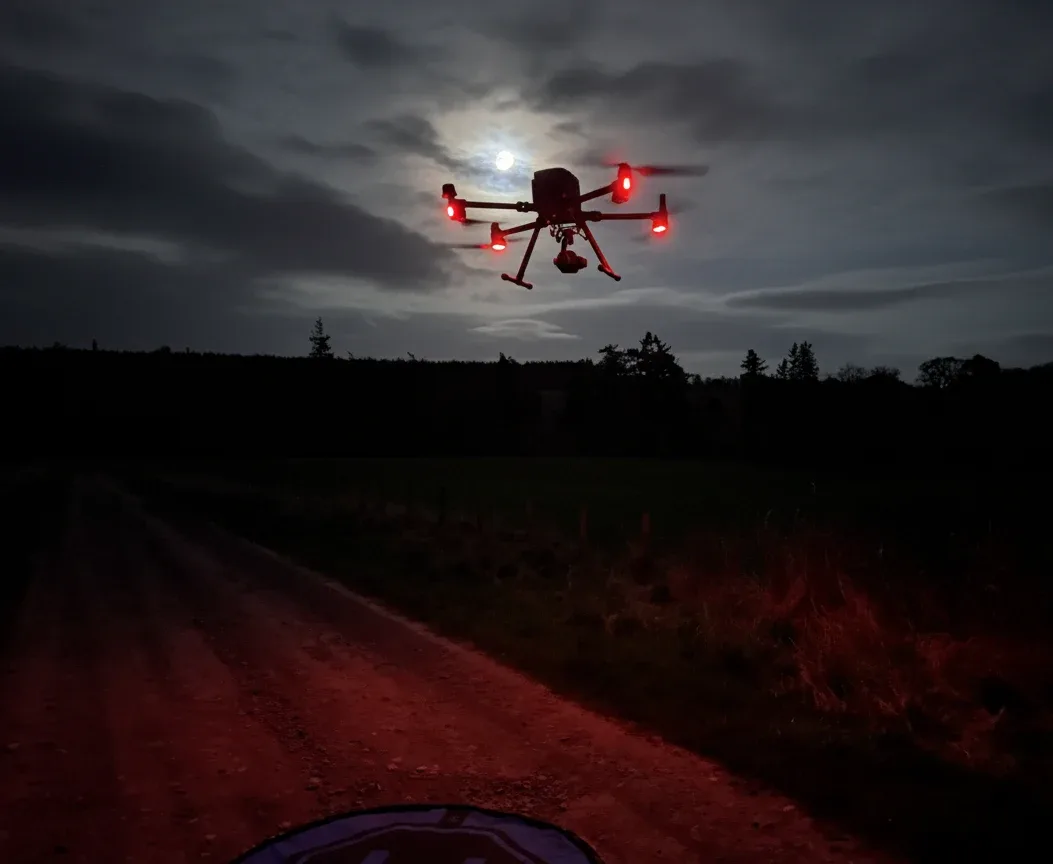 A large drone hovering in front of the moon at dusk over a farm track, its arm lights glowing red rather than green