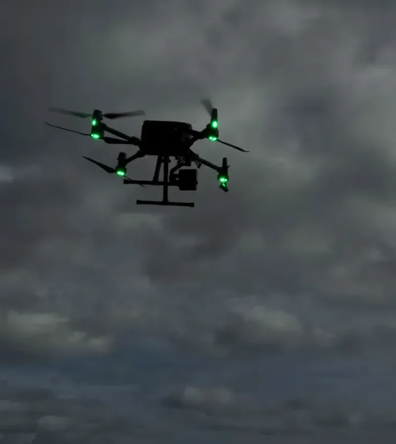 A drone hovering against heavy grey cloud at dusk with four bright green lights glowing on the ends of its arms