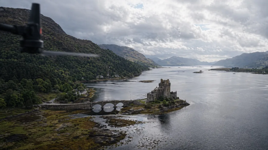Aerial photograph of Eilean Donan castle and its arched bridge on a Scottish loch, taken from out over open water with hills behind, a drone propeller blurred in the top corner