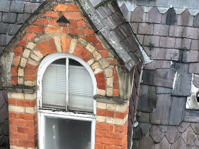 Drone photograph of a brick dormer showing perished mortar around the arch, moss on the slate courses and a repaired patch of dark render on the roof beside it