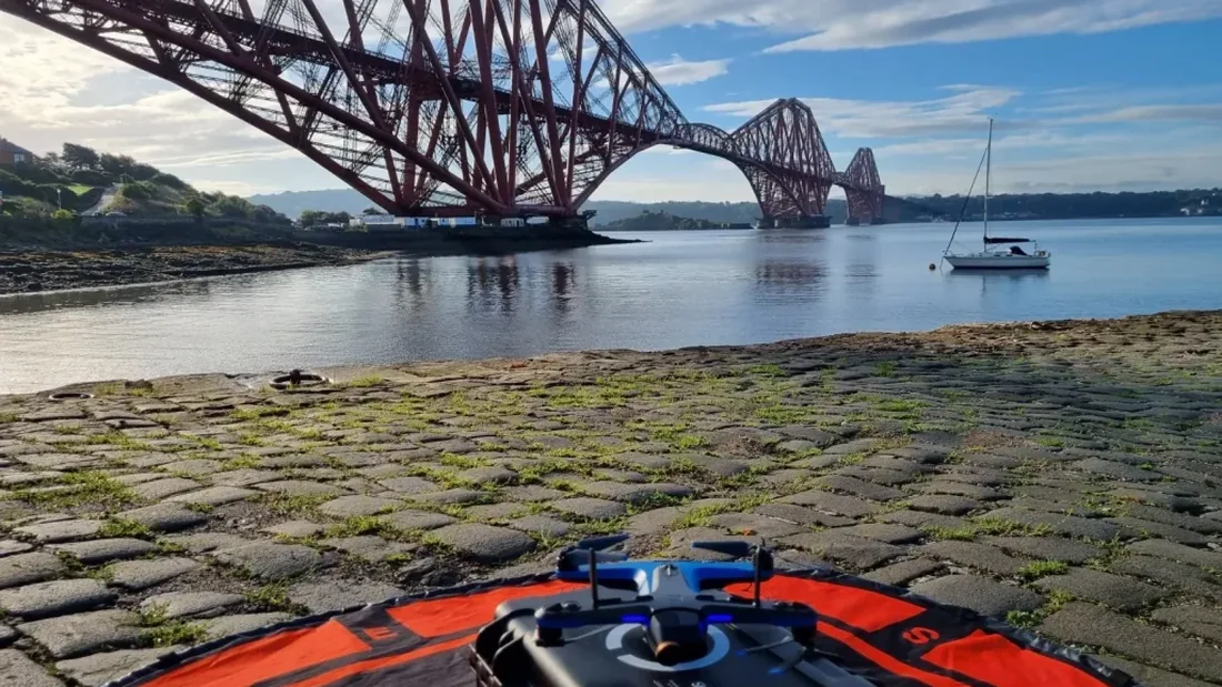 A small drone sitting on an orange landing pad on a stone jetty, with the Forth Bridge and open water behind it