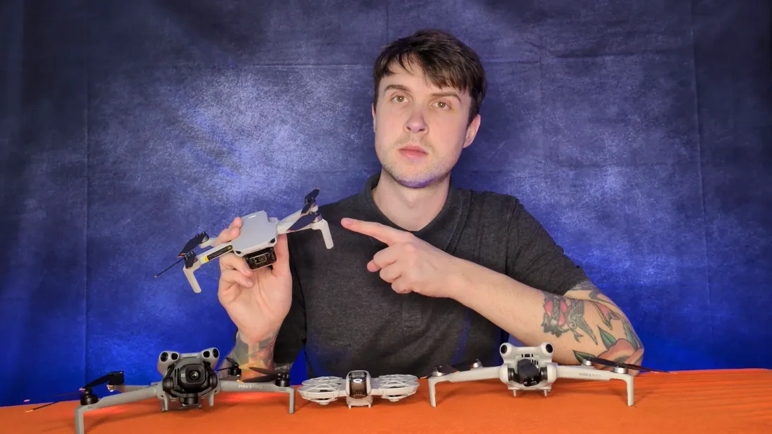 Peter Leslie holding up a small folding DJI drone with a DJI Mini 5 Pro, a caged DJI Neo and a DJI Mini 4 Pro lined up on the desk in front of him