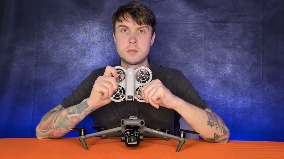 Peter Leslie holding a small caged DJI Neo 2 in both hands above a much larger DJI Mavic 3 Pro sitting on the desk, showing the difference in mass between two drones rated close together for wind