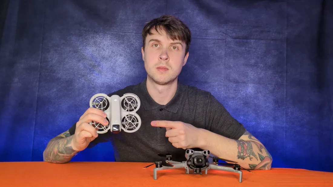 Peter Leslie holding up a caged DJI Neo 2 and pointing at it, with a DJI Mini 5 Pro on the desk in front of him