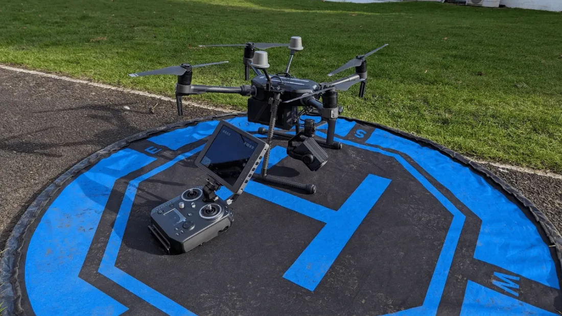 A drone and its screen controller set out on a marked landing pad on tarmac in low winter sunlight, ready for take-off
