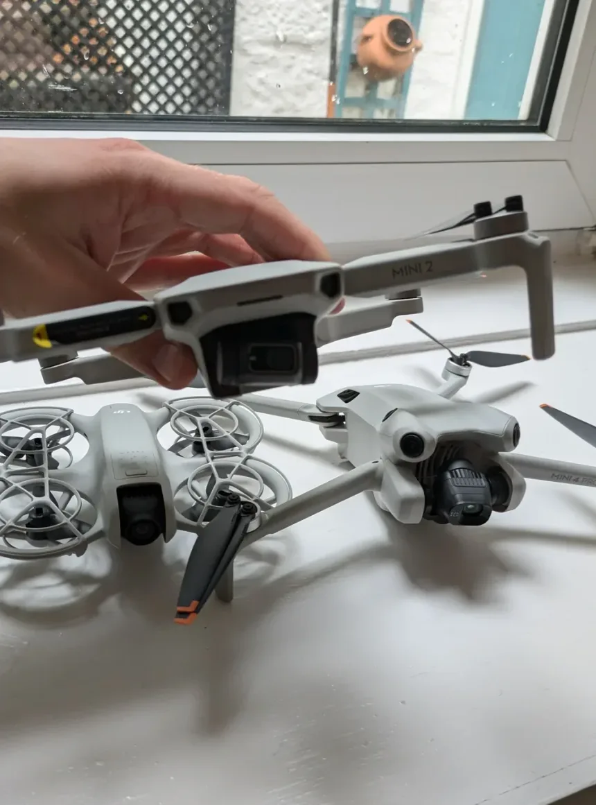 Three small DJI drones on a white windowsill — a caged DJI Neo, a folded DJI Mini 2 held in one hand, and a DJI Mini 4 Pro with its gimbal camera exposed