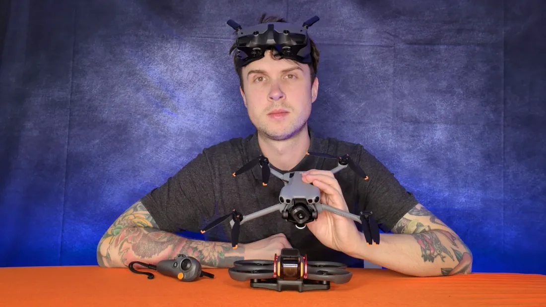 Peter Leslie wearing DJI FPV goggles pushed up on his forehead, holding a folding camera drone, with an FPV drone and a motion controller on the table in front of him