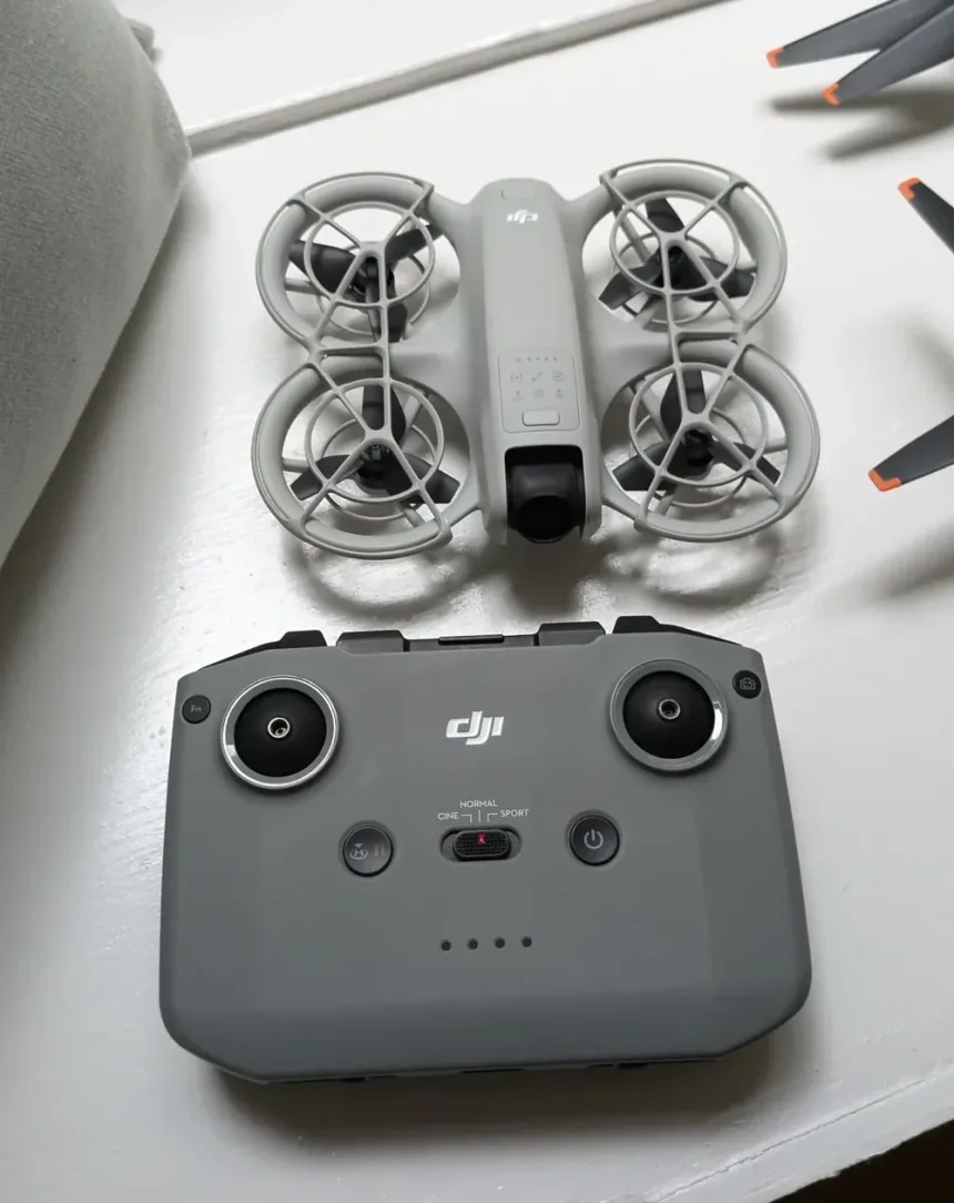 A caged DJI Neo drone lying on a white surface next to a grey DJI RC-N3 remote controller with its control sticks fitted