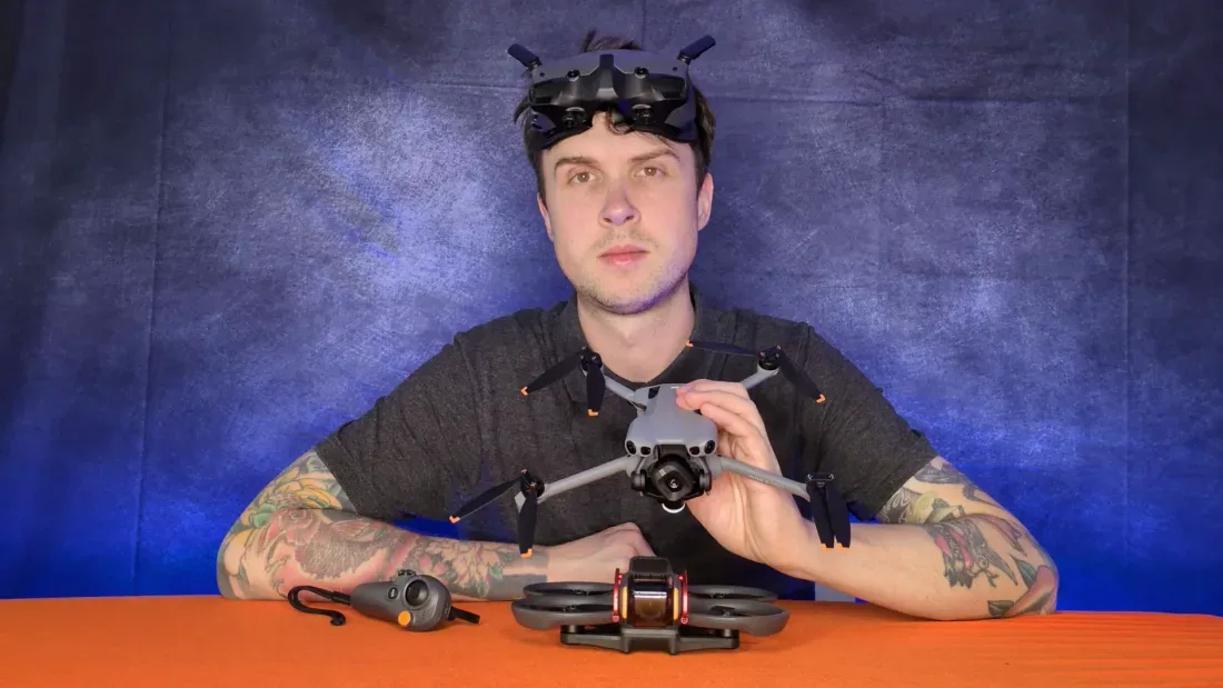 Peter Leslie with DJI Goggles 3 resting on his forehead, a DJI Mini in his hands and a DJI Avata 2 with an RC Motion controller laid out on the desk