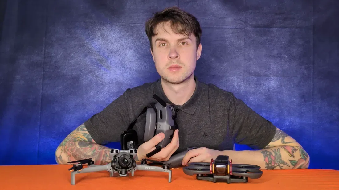 Peter Leslie holding a DJI RC-N controller with a folded DJI Mini 4 Pro on the desk in front of him and a DJI Avata FPV drone beside it