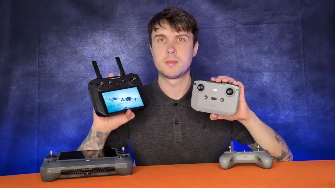 Peter Leslie holding up two drone controllers, a screen-based professional controller and a phone-mount controller, with two more on the table in front of him