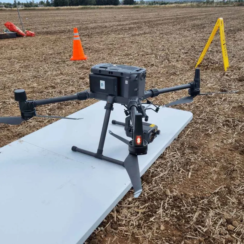 An enterprise survey drone with a sensor payload sitting on a white landing pad in a ploughed field, with a traffic cone and a folding warning sign marking the take-off area